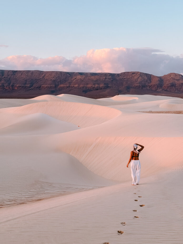 A woman walks barefoot across the sweeping golden sand dunes of Socotra at sunset, leaving footprints toward rugged mountains—one of the most unforgettable things to do in Socotra for travelers seeking beautiful, otherworldly landscapes beyond the tourist trail.