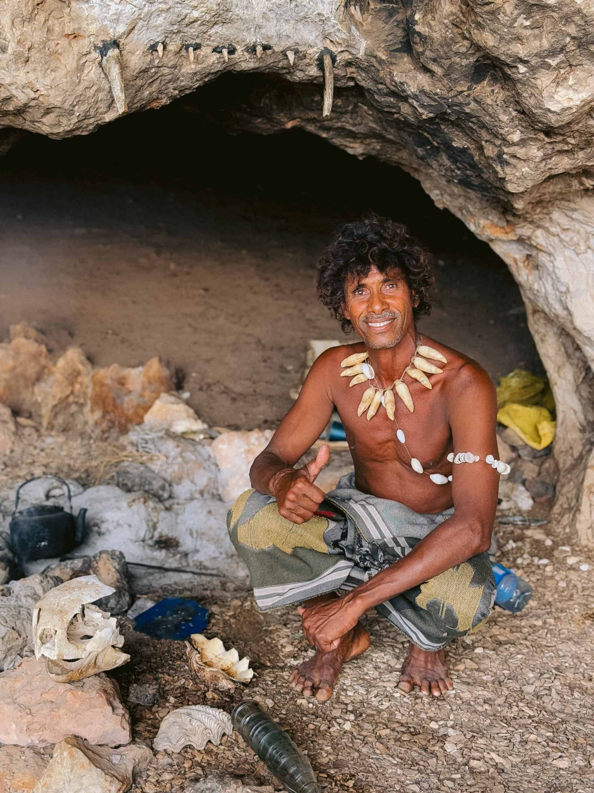 Socotra's cave man smiling at the entrance of a cave, wearing a necklace made of animal teeth and shells, surrounded by natural artifacts and traditional items — a unique cultural encounter and one of the memorable things to do in Socotra.