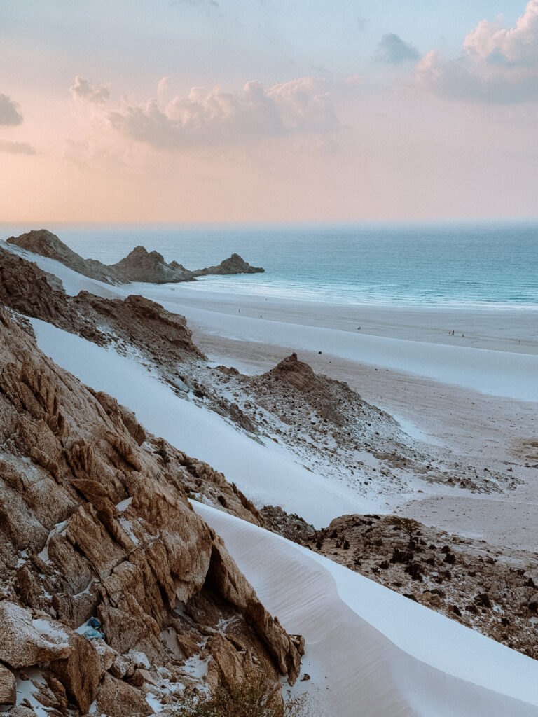 White sand dunes meeting turquoise waters at Qalansiyah Beach in Socotra, framed by rugged rocky cliffs at sunset, showcasing the island’s unique coastal landscapes.
