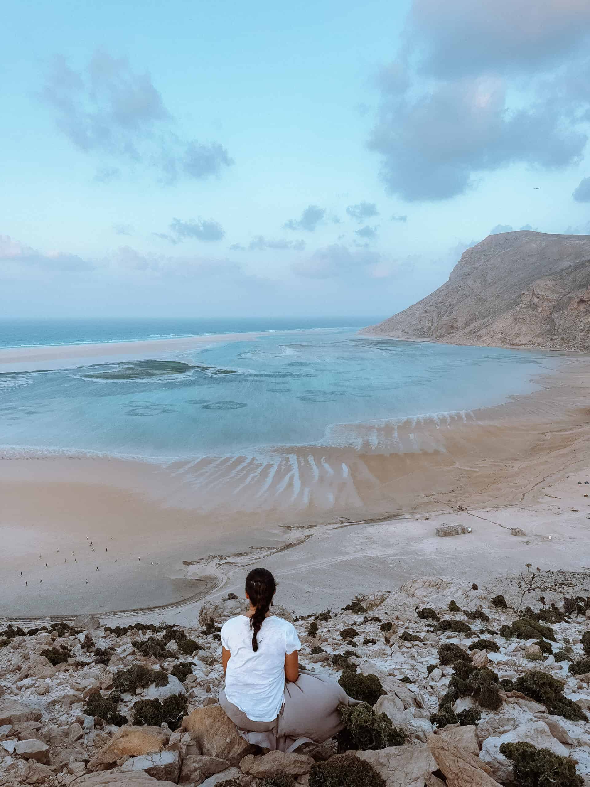 A woman sits on rocky terrain overlooking the expansive turquoise and sandy tidal flats of Detwah Lagoon in Socotra at low tide, framed by rugged mountains on the right and the Arabian Sea on the horizon under a partly cloudy sky — one of the most breathtaking things to do in Socotra.