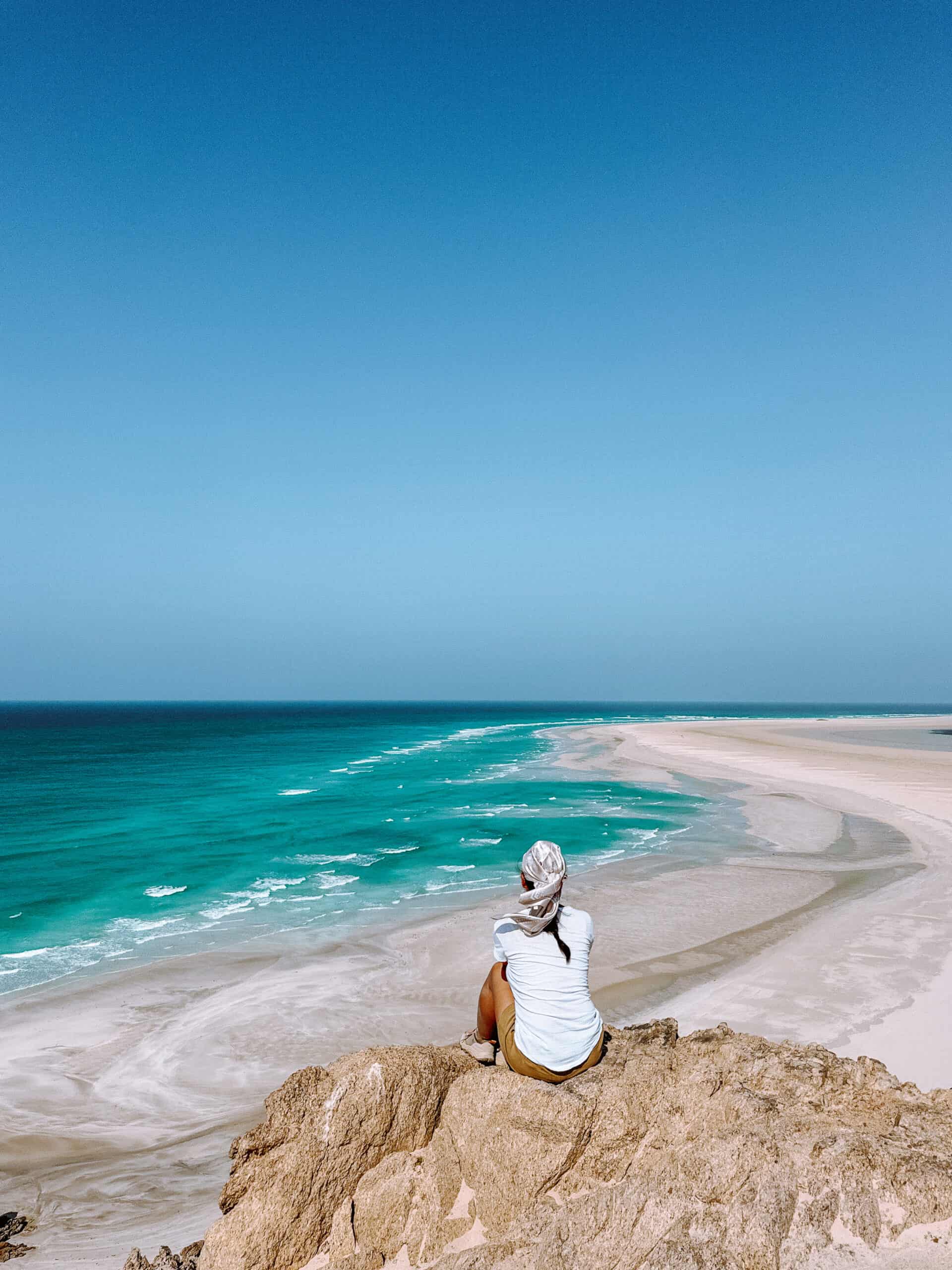 A traveler sits on a rocky cliff at one of Socotra's stunning beaches, gazing at the sweeping turquoise waters and sandbars of Socotra Island’s coastline — one of the most breathtaking viewpoints and must-see spots when exploring the best things to do in Socotra.