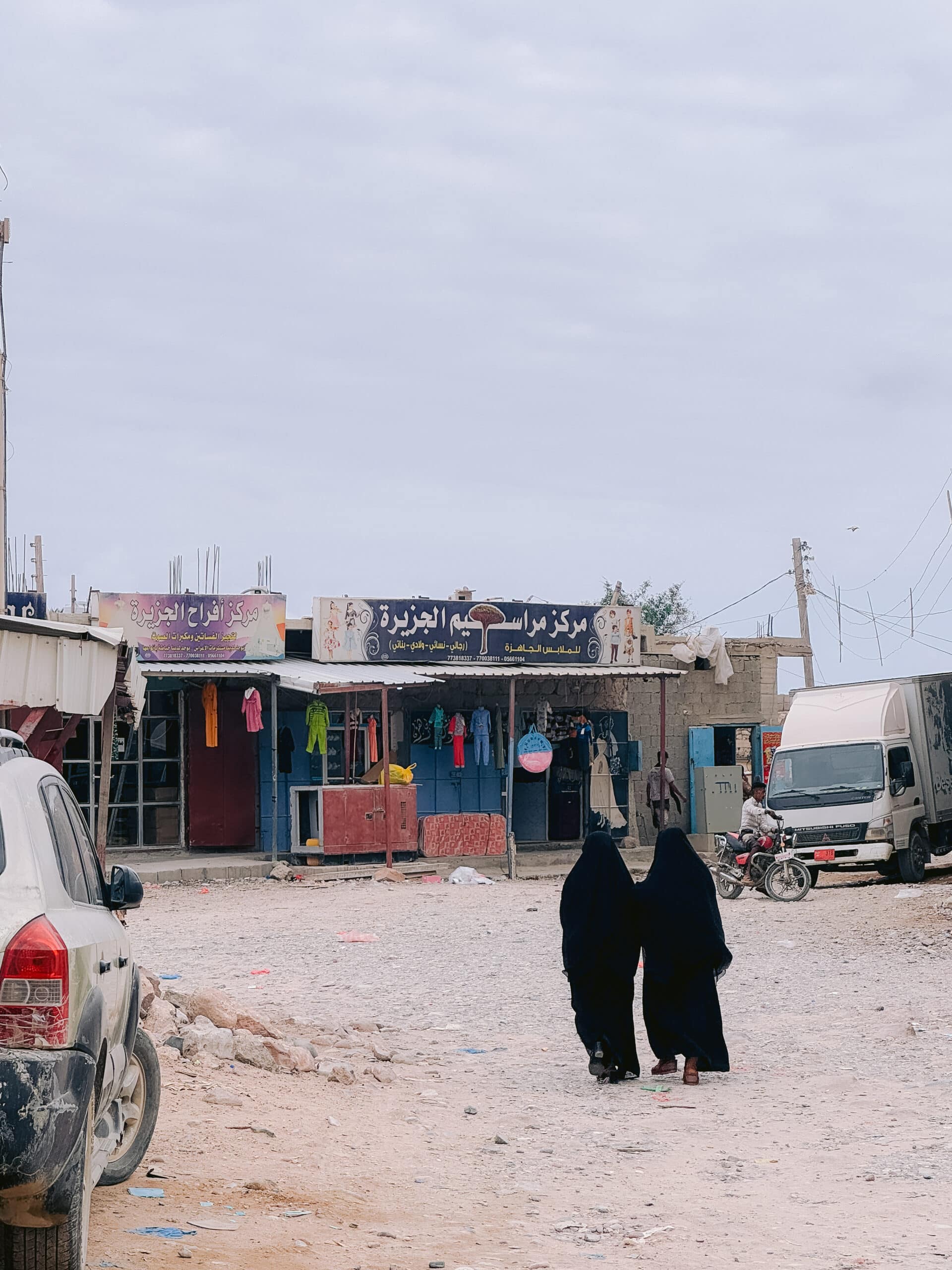 Two women dressed in black abayas walk down a dusty, unpaved street in Hadibo, the capital of Socotra, Yemen, with small local shops, colorful clothes hanging outside, and vehicles parked along the roadside under a cloudy sky.