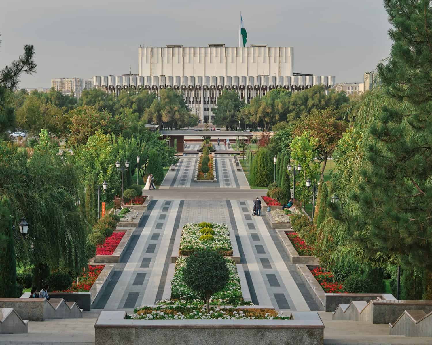 Lush city park in Tashkent with well-maintained flower beds, symmetrical pathways, and mature trees leading to a modern government building in the background. Perfect for travel and urban exploration.