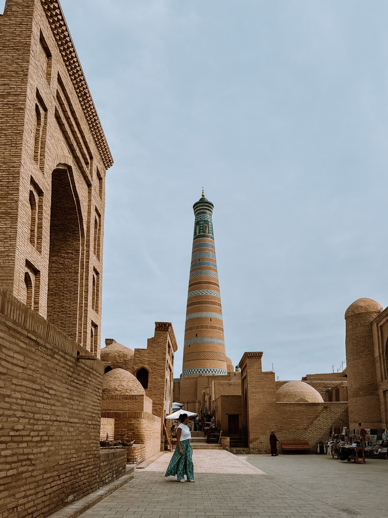 Traveler walking down a sunlit street toward the Islam Khodja Minaret in Khiva’s old town — a classic Silk Road scene.