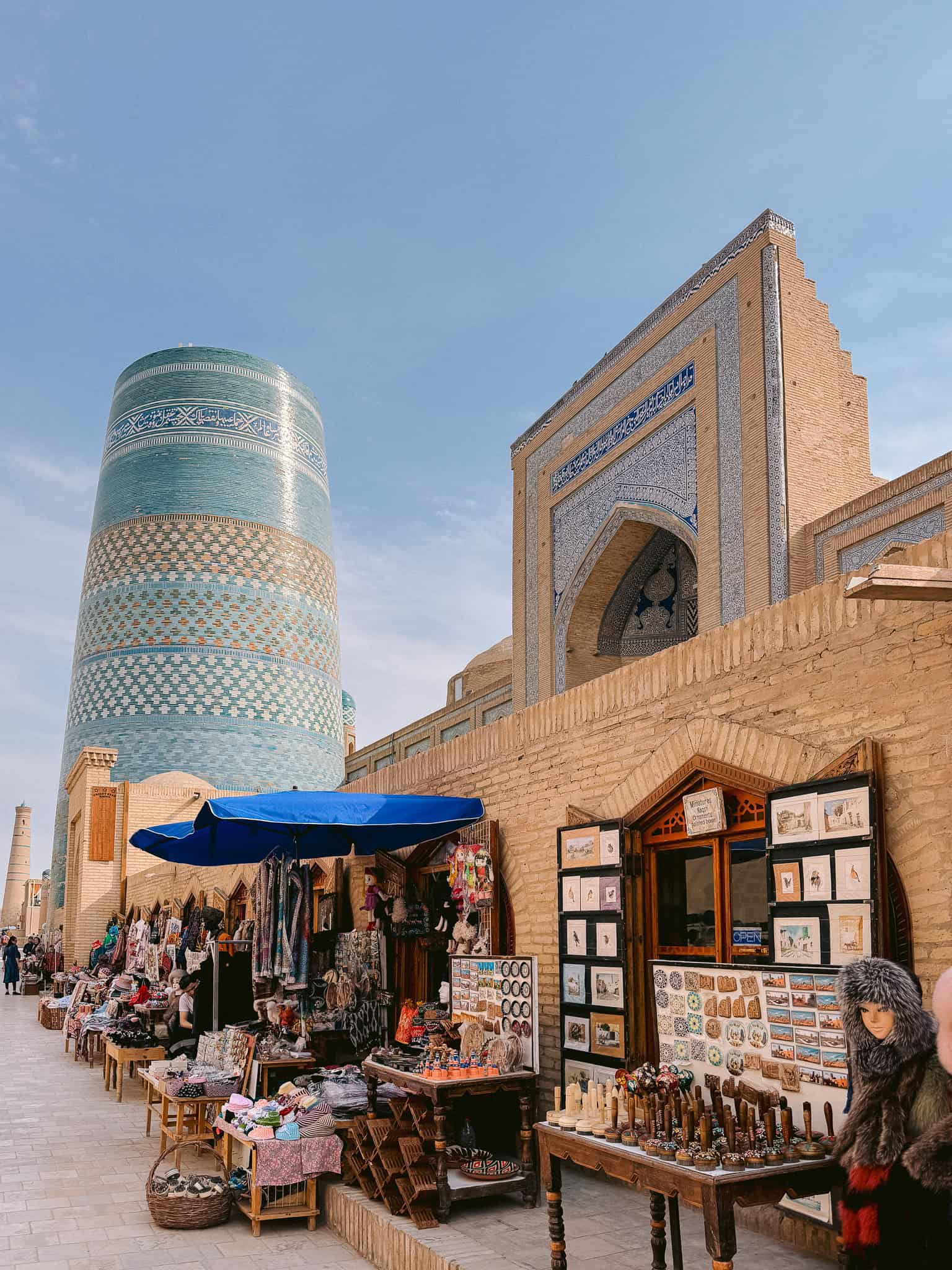 Vendors and souvenir shops line the street near Khiva’s turquoise Kalta Minor Minaret — a lively corner of the old walled city.
