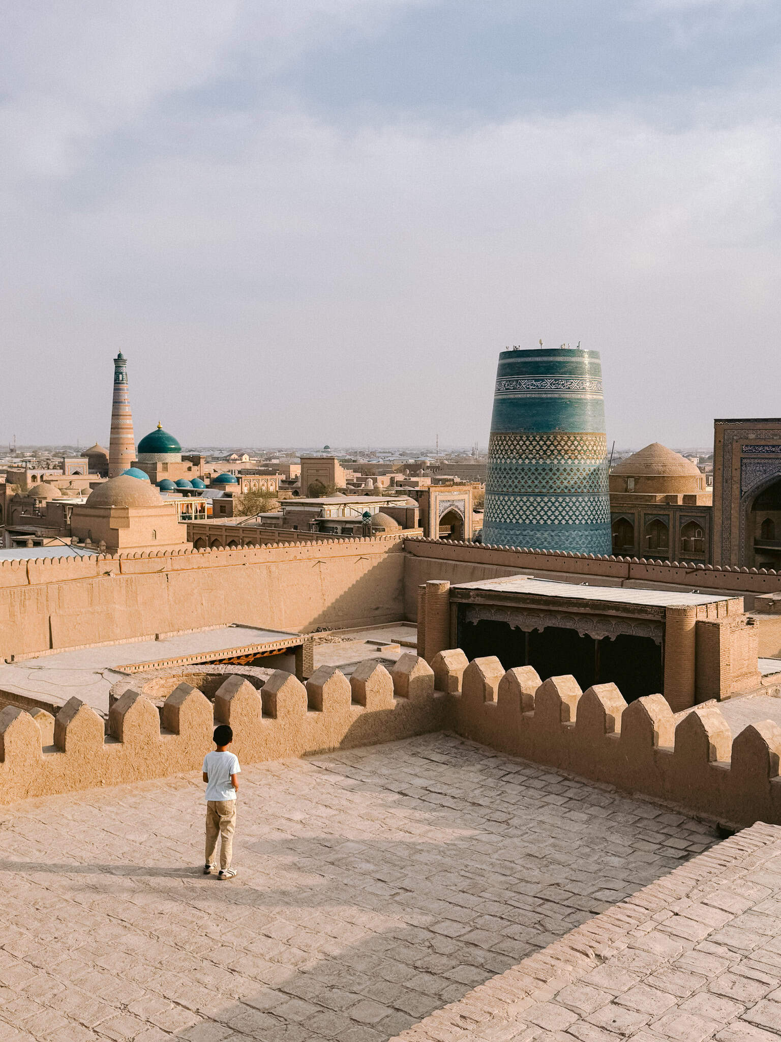 Boy standing on the ramparts of Khiva’s Kuhna Ark fortress with panoramic views over Itchan Kala’s minarets and domes.